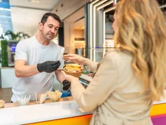 Food vendor wearing black gloves handing over a personalized fruit bowl with granola and various fruits to a female customer in a modern shopping mall food stall. Food price caps.
