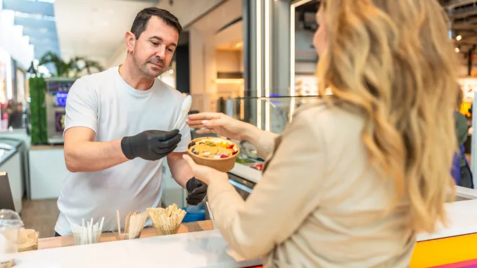 Food vendor wearing black gloves handing over a personalized fruit bowl with granola and various fruits to a female customer in a modern shopping mall food stall. Food price caps.